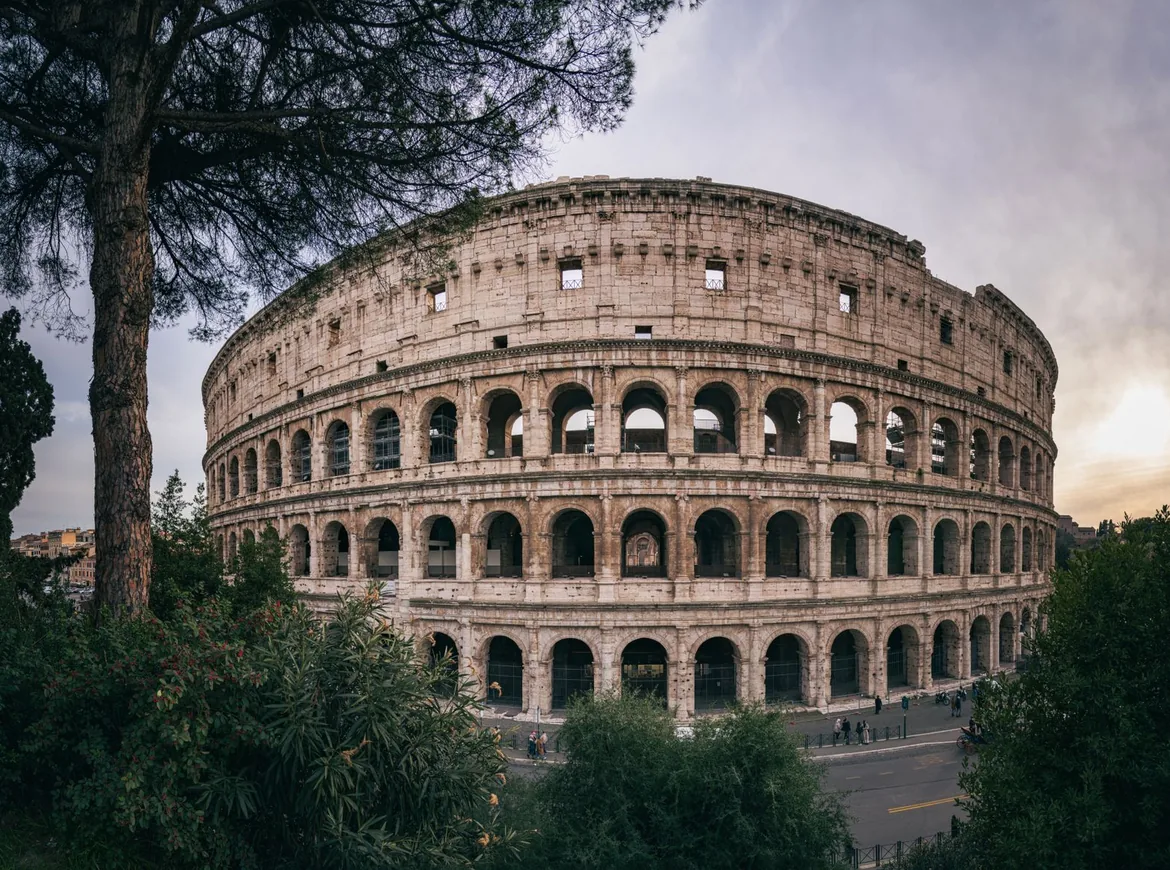 Colosseum i Rom under en blød aftenhimmel, indrammet af pinjetræer