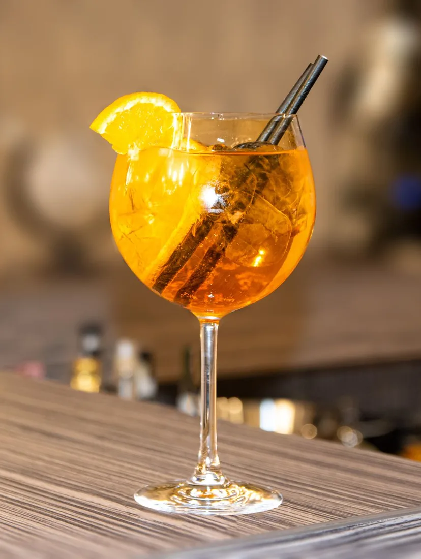Et højt glas orange spritz med en appelsinskive og et sugerør på en træbar