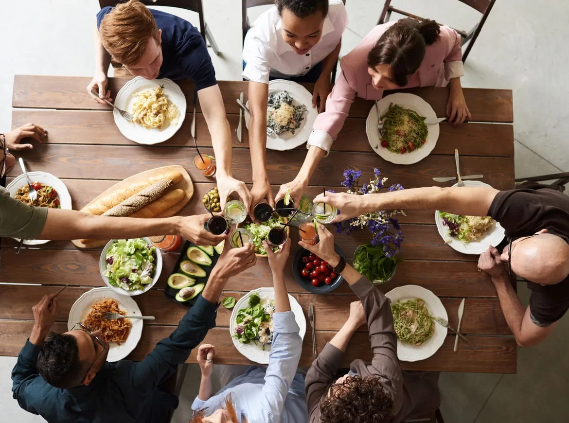 Set ovenfra: venner ved et træbord, der skåler over tallerkener med pasta og salat